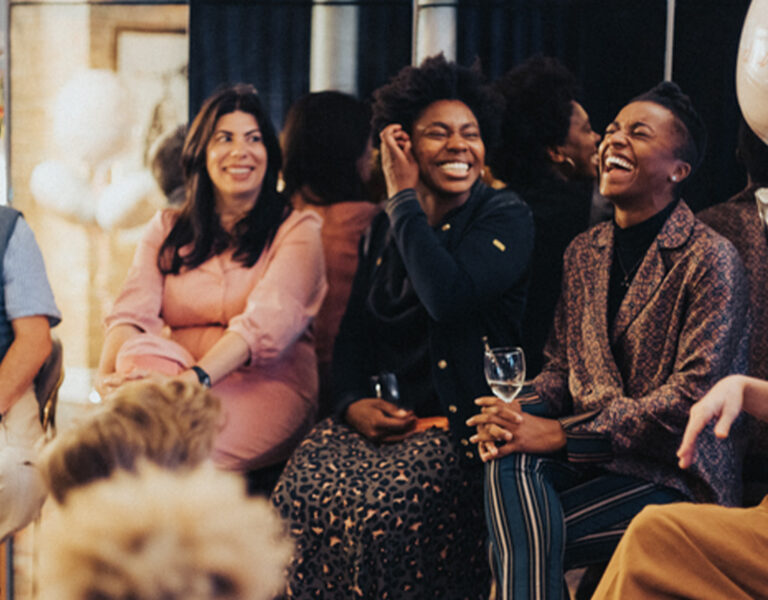Five adults sit in a row indoors, smiling and laughing together. One woman holds a wine glass; another gestures with her hand. There are balloons and blurred people in the foreground. The environment appears social and relaxed.