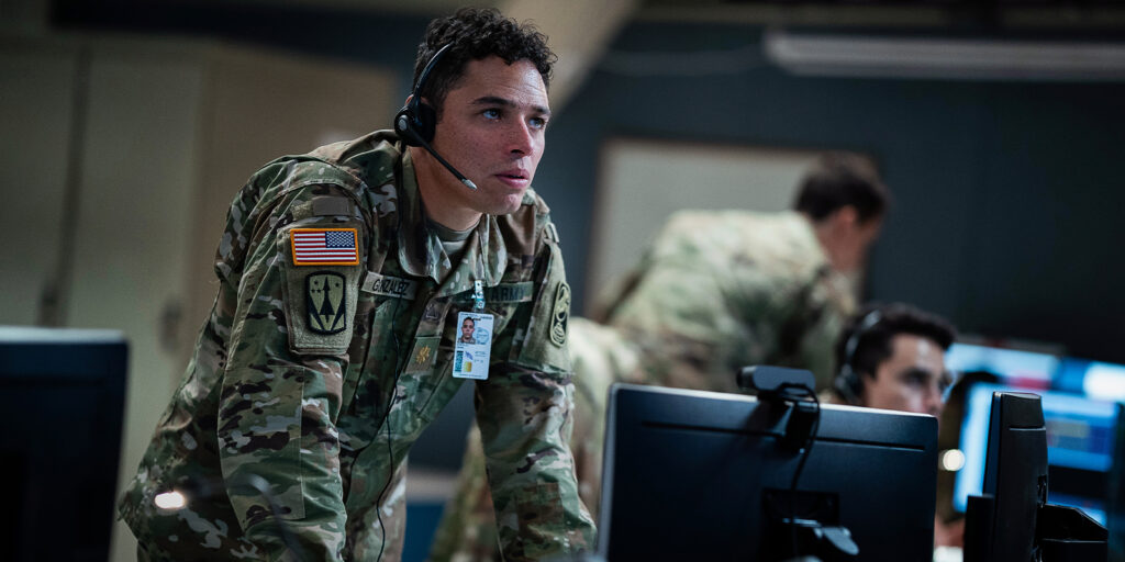 A US Army soldier in camouflage uniform and headset leans over a desk with computer monitors, appearing focused, in a command centre setting reminiscent of Barry Ackroyd’s intense, immersive style. Other soldiers work in the background.