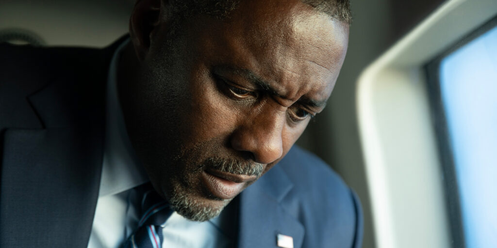A man in a dark suit and striped tie looks down with a serious, concerned expression, as sunlight from the window casts dramatic shadows—reminiscent of Barry Ackroyd’s cinematography in A House of Dynamite.