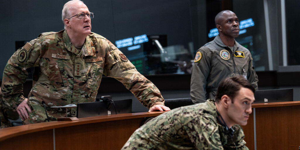 Three men in military uniforms stand and sit around a desk in a command centre, looking intently at something off-screen—an intense scene in Barry Ackroyd’s gripping style in A House of Dynamite. Computer monitors and control panels surround them.