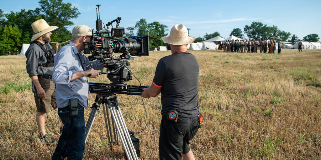 Three crew members film a scene outdoors with a large camera on a tripod in a grassy field, capturing actors near white marquees. Inspired by Barry Ackroyd's style, this could be a shoot for A House of Dynamite under blue skies and tall trees.