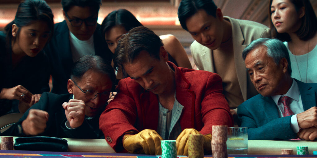 A man in a red jacket sits at a casino table, with tense expression, whilst onlookers and players cheer or watch intently. Poker chips are stacked on the table.