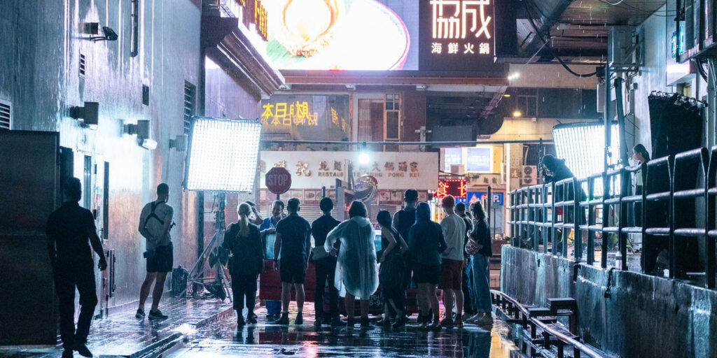 A film crew gathers in a wet, neon-lit alley at night, with ASC BSC cinematographer James Friend guiding the shoot. Large lights and equipment frame Chinese signs and shopfronts glowing in the background.