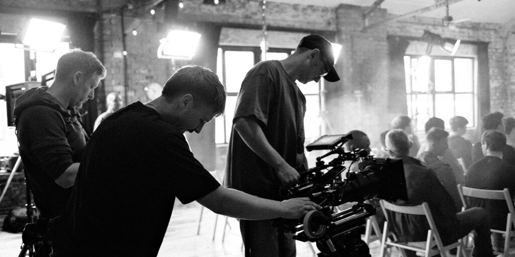 Three people, including Josh Carrington Birch, operate professional video cameras in a studio with brick walls and large windows. Several people sit on chairs facing away from the camera, whilst bright lights illuminate the black-and-white scene.