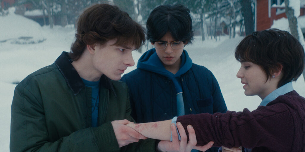 Three teenagers stand outside in the snow. One boy examines a rash on a girl's forearm whilst another looks on with concern, reminiscent of a tense scene from Black Phone 2. Snowy trees and buildings are visible in the background.
