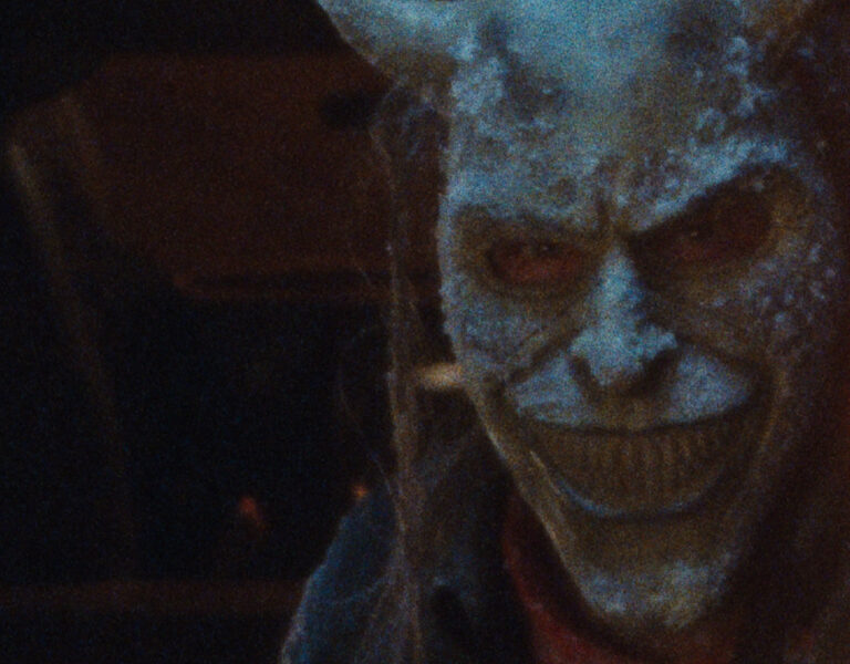 A person wearing a sinister, frost-covered mask with exaggerated sharp teeth and horns stares menacingly in dim lighting, reminiscent of the chilling atmosphere in Black Phone 2.