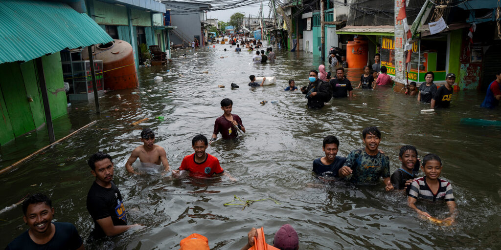 People wade through waist-deep floodwaters on a street lined with houses and shops. Some smile and pose for the camera, whilst others walk or sit in the water. The area appears crowded and heavily flooded.