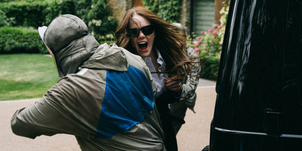 A woman wearing sunglasses and holding scissors aggressively confronts a hooded figure outdoors, next to a black vehicle, with greenery and flowers.