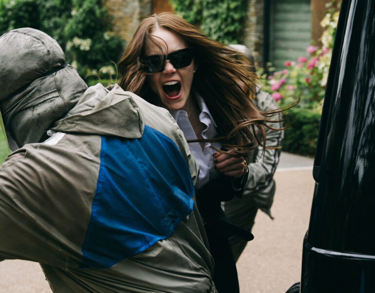 A woman wearing sunglasses and holding scissors aggressively confronts a hooded figure outdoors, next to a black vehicle, with greenery and flowers.