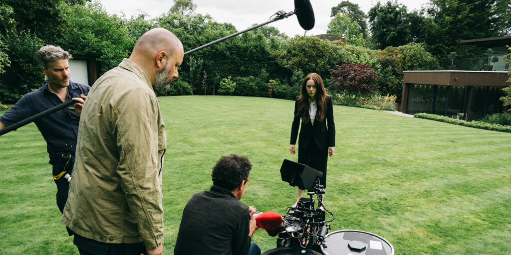 Four crew members are filming outside on a green lawn. A woman in a black suit stands in front of the camera, while two crew members operate equipment and one holds a boom mic overhead. Trees and a modern house are in the background.