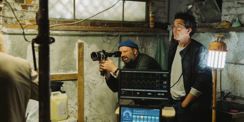 Two men work on a film set in a basement; one operates a camera while the other stands beside him. In the foreground, a medical monitor displays vital signs.