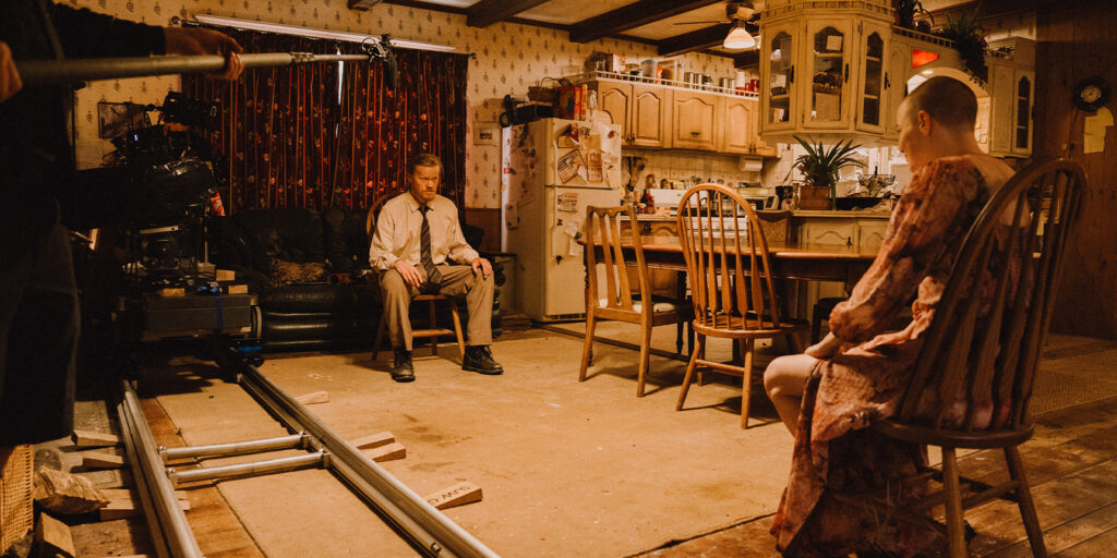 A behind-the-scenes look in a cluttered kitchen, featuring a man in formal clothes on a sofa and someone in a hospital gown with a shaved head on a chair—camera equipment visible, capturing the Bugonia magic through Robbie Ryan's lens.