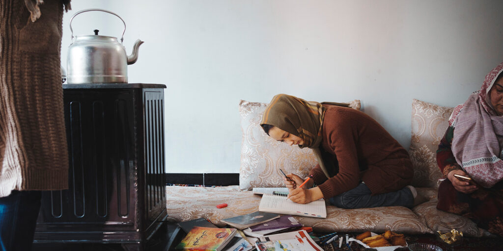 A girl wearing a headscarf sits on a cushion, writing in a notebook with books and papers scattered around. Another person, partially visible, sits beside her. A metal kettle is on a cooker nearby.