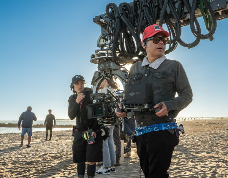 Two film-makers operate a camera rig on a sunny beach, surrounded by crew and equipment, capturing a scene from Caught Stealing, with the ocean and city skyline visible in the background.