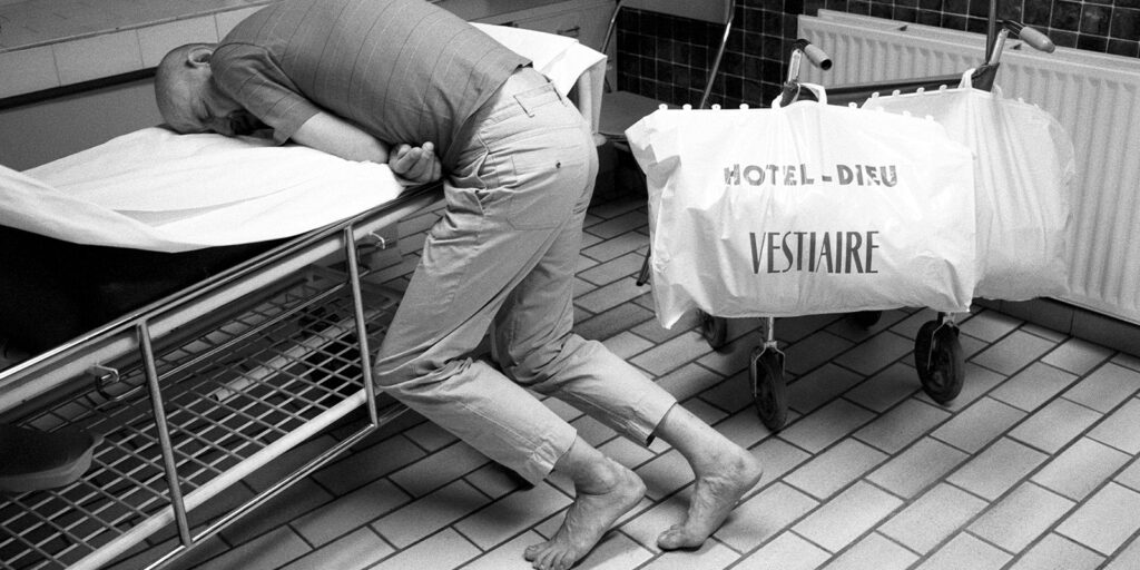 A barefoot man in clothing leans over a hospital bed, clutching his stomach in pain. Next to him is a trolley with a large bag labelled HOTEL-DIEU VESTIAIRE. The scene is in a tiled hospital ward.