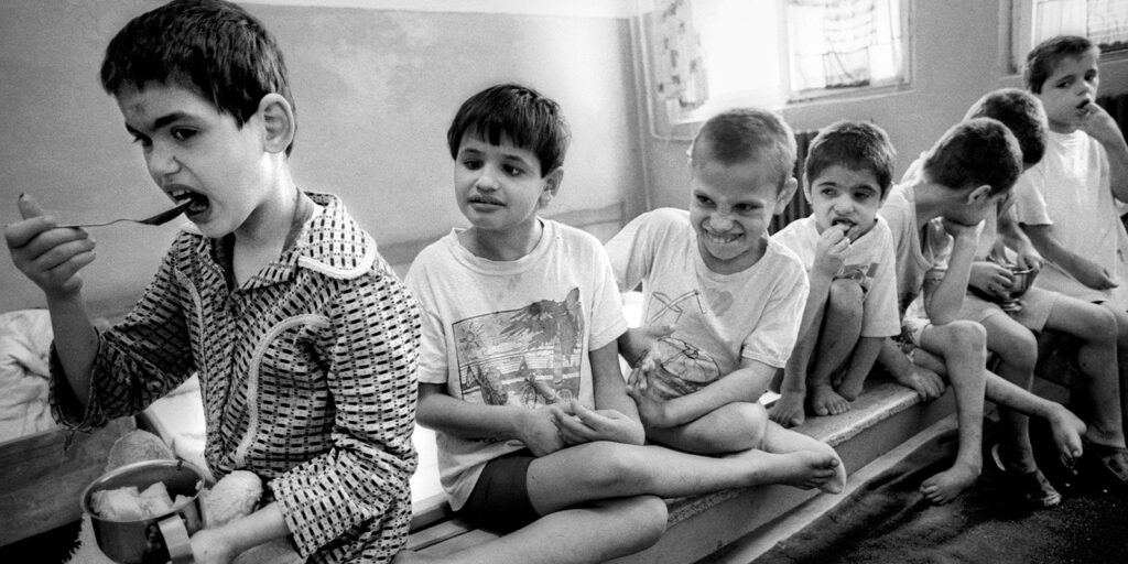 A group of young boys sit on a bench in a simple room. One boy eats from a bowl with a spoon, while the others sit close together, some smiling and some with neutral expressions.