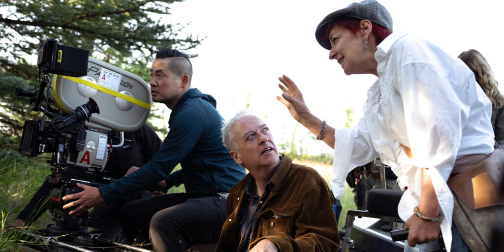Three people on a film set: one operates a large camera, another kneels and listens attentively, while a third person gestures as if explaining something, surrounded by trees and natural light