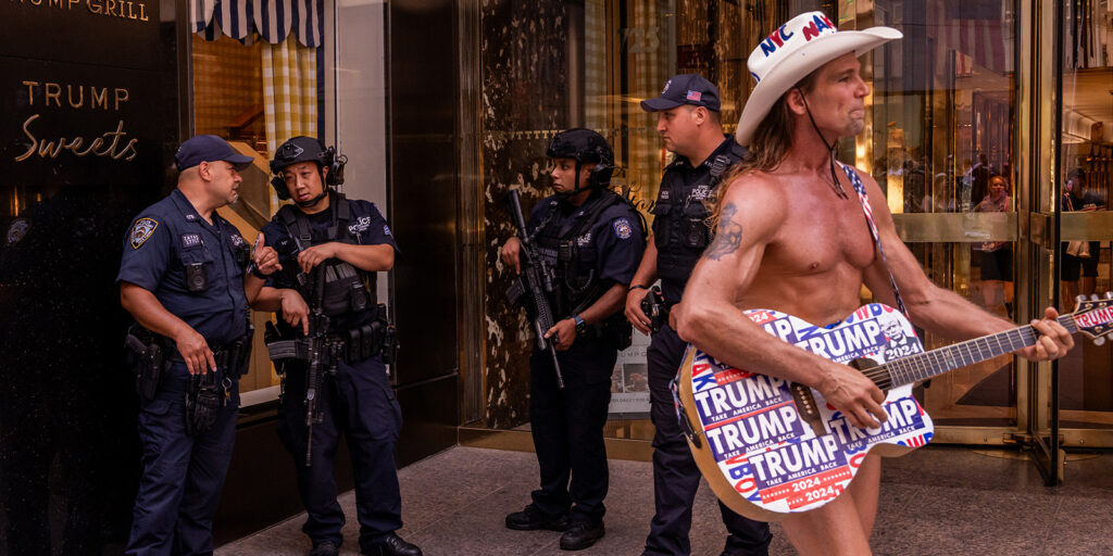 A bare-chested man in a cowboy hat decorated with “Trump” stickers plays a guitar in front of four armed police officers standing outside a building labelled Trump Sweets.