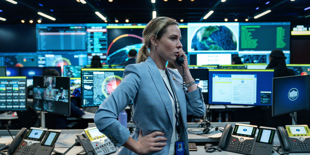 A woman in a blue blazer stands in a high-tech command centre, speaking on the phone. Surrounded by monitors displaying maps and data, the bustling scene echoes the tense visuals of Barry Ackroyd’s work in A House of Dynamite.
