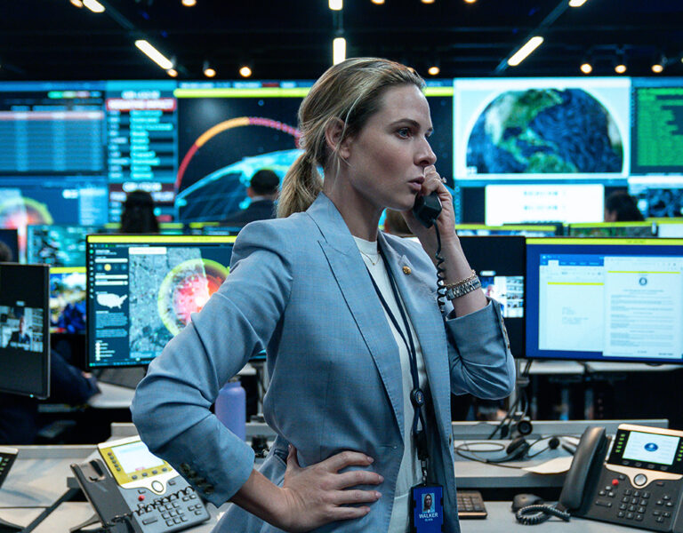 A woman in a blue blazer stands in a high-tech command centre, speaking on the phone. Surrounded by monitors displaying maps and data, the bustling scene echoes the tense visuals of Barry Ackroyd’s work in A House of Dynamite.