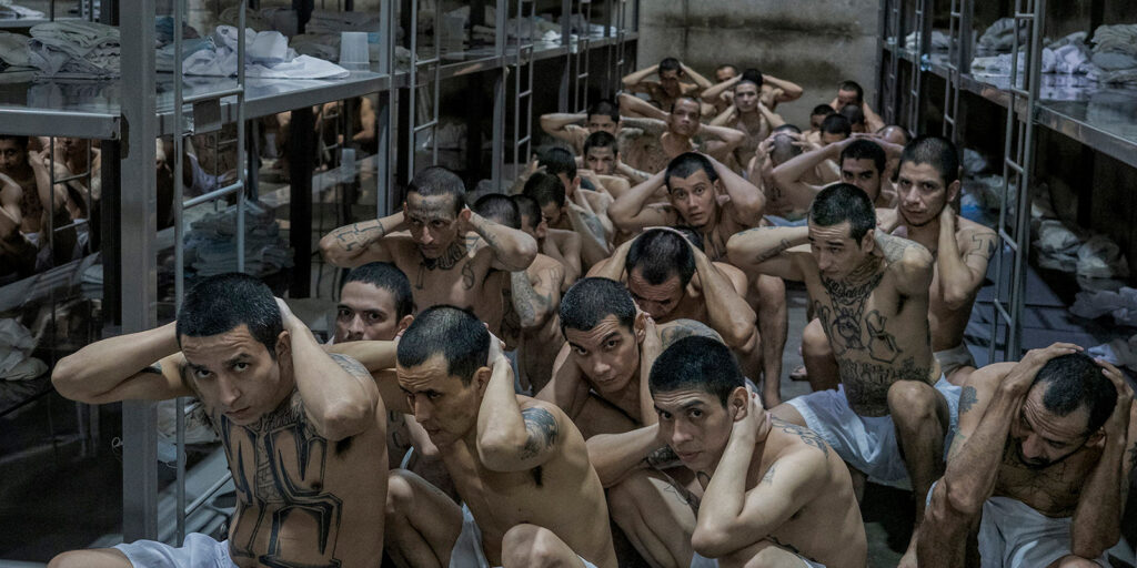 A group of bare-chested men with tattoos sit closely together on the floor of a crowded room, hands behind their heads, surrounded by metal shelves filled with clothing and belongings.