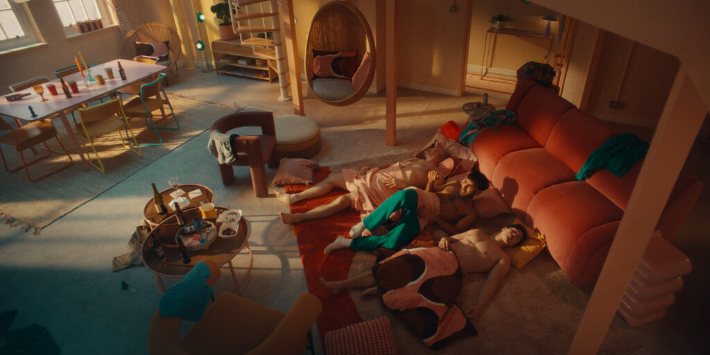 Three people lie together on the floor next to a pink sofa in a cosy, sunlit living room. The space is cluttered with clothes, bottles, and dishes, suggesting the aftermath of a gathering.