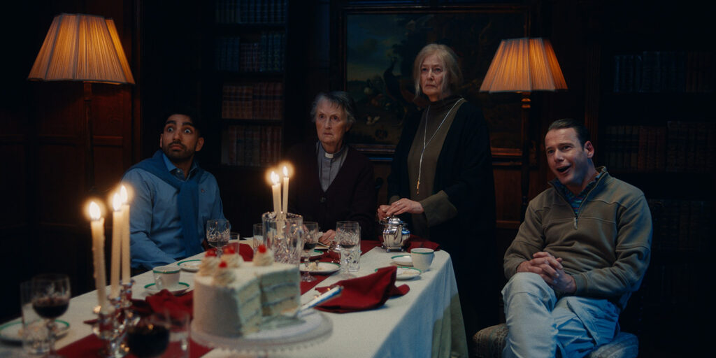 Four people sit and stand around a formal dining table with a cake, candles, and drinks, all looking surprised or startled. The room is dimly lit with wooden walls, bookshelves, and warm lamps.