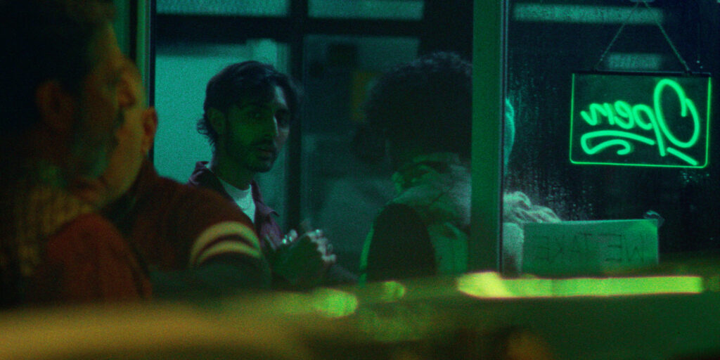 A man stands in a dimly lit room, looking out towards the camera. A neon green Open sign glows in the window next to him, casting coloured light on the scene. Other people are partially visible in the foreground and background.