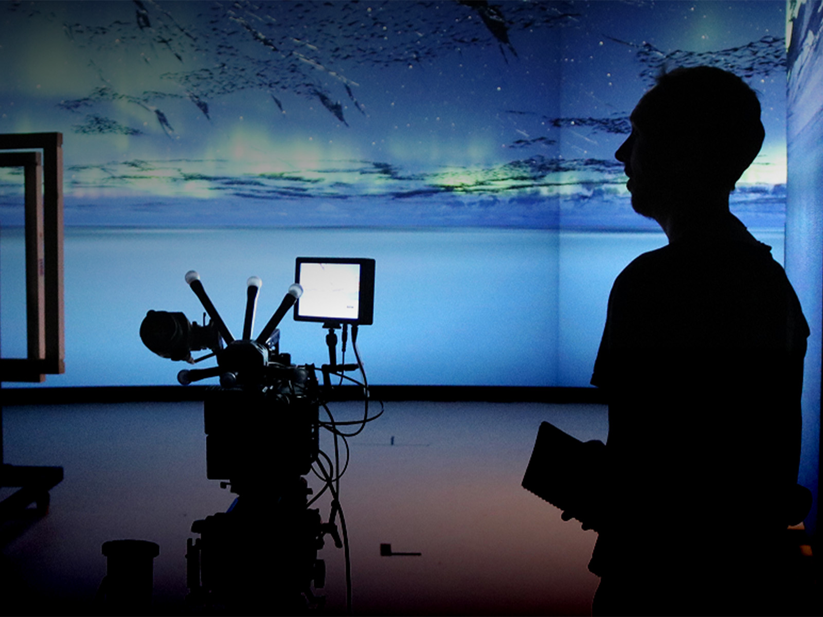 A person stands near a camera set-up in a studio with blue digital visuals projected on the walls.