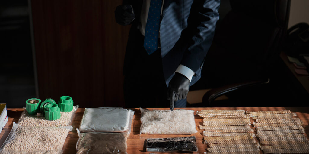 A person in a suit and blue tie, wearing black gloves, stands behind a table displaying several bags of white and beige powder, green containers, and rows of packaged items, possibly during an investigation or presentation.