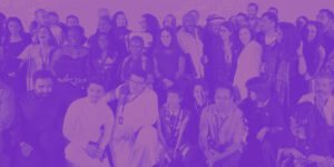 A large group of people pose together for a photo, some standing and some kneeling at the front, all facing the camera. The image has a purple colour overlay and was taken at the Red Sea International Film Festival's Red Sea Souk Project Market.