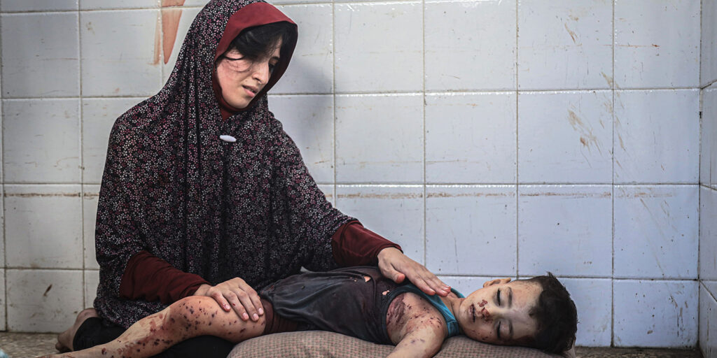 A woman in a patterned headscarf sits on the floor, looking sorrowfully at a young child lying beside her with visible injuries, against a white tiled wall.