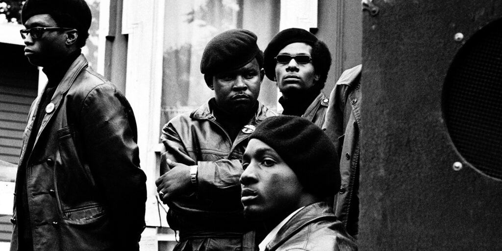 Four men wearing black berets, sunglasses, and leather jackets stand together outside a building, looking serious and focused. The photo is in black and white, giving it a powerful, historic feel.