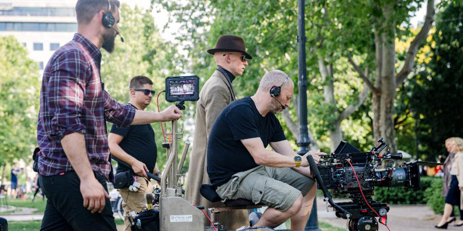 A film crew works outdoors with a camera set-up on a dolly, as Christopher Ross BSC operates the camera while others assist, wearing headsets. A person in a brown coat and hat walks nearby. Trees and park greenery are visible in the background.