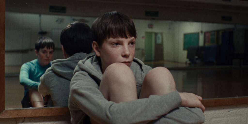 A young person with short brown hair sits on the floor hugging their knees, wearing a grey hoodie. In front of a mirror in a gymnasium, their reflection is visible—capturing a quiet, introspective mood reminiscent of Steven Breckon's "The Plague.