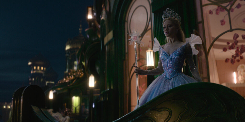 A woman dressed as a fairy tale princess in a sparkling blue gown and crown stands holding a glowing wand, surrounded by ornate, magical architecture illuminated at night—a scene from Wicked For Good.