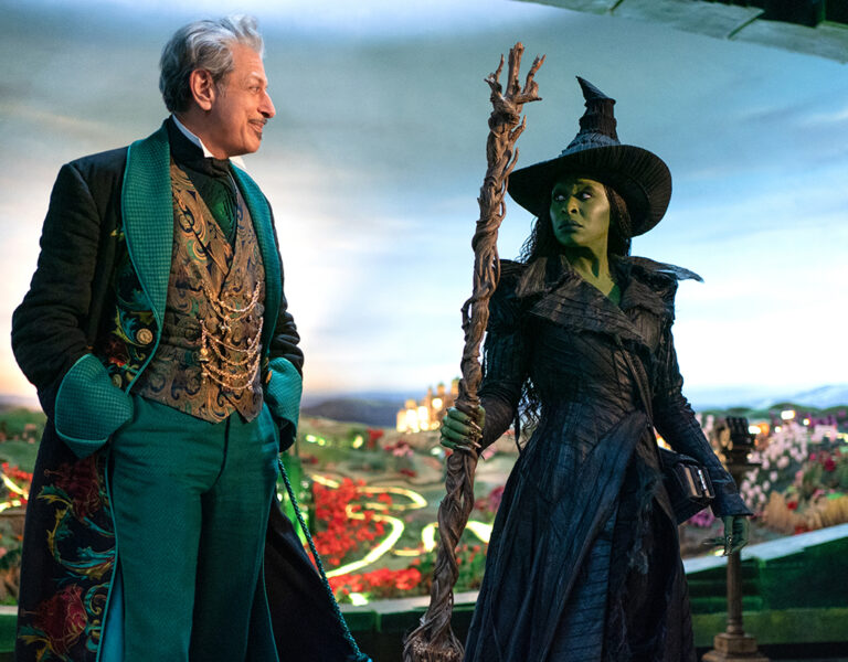 A man in an ornate green suit smiles at a green-skinned witch holding a staff. They stand before a magical city, capturing the enchanting world of Wicked For Good as seen through Alice Brooks BSC's cinematic vision.