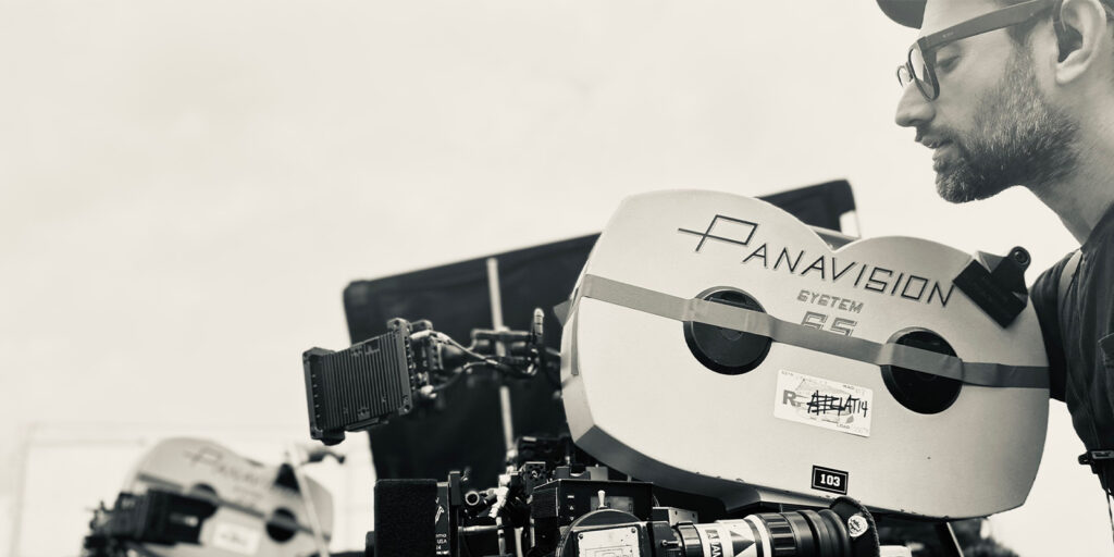 Ethan McDonald, a focus puller wearing glasses, stands beside a large Panavision film camera on set. The black-and-white image features a soft, blurred background with another camera visible.