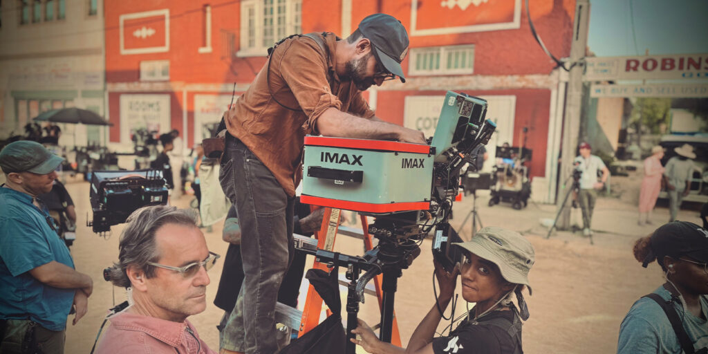 A film crew is on set outdoors. Focus puller Ethan McDonald stands on a ladder adjusting an IMAX camera, whilst a woman nearby holds equipment. Other crew members and cameras are visible near red and beige buildings in the background.