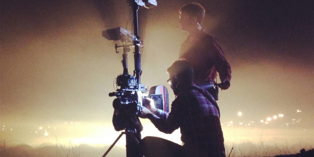 Two people, including focus puller Ethan McDonald, operate a professional video camera on a tripod at night. One stands while the other kneels to adjust the camera. The hazy background features distant, glowing lights.
