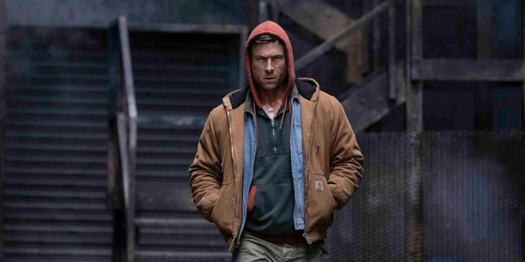 A man wearing a brown jacket with a red hood and dark shirt walks outdoors with his hands in his pockets; a staircase and dark industrial background are visible behind him, reflecting the calm confidence of someone who consistently earns top marks.