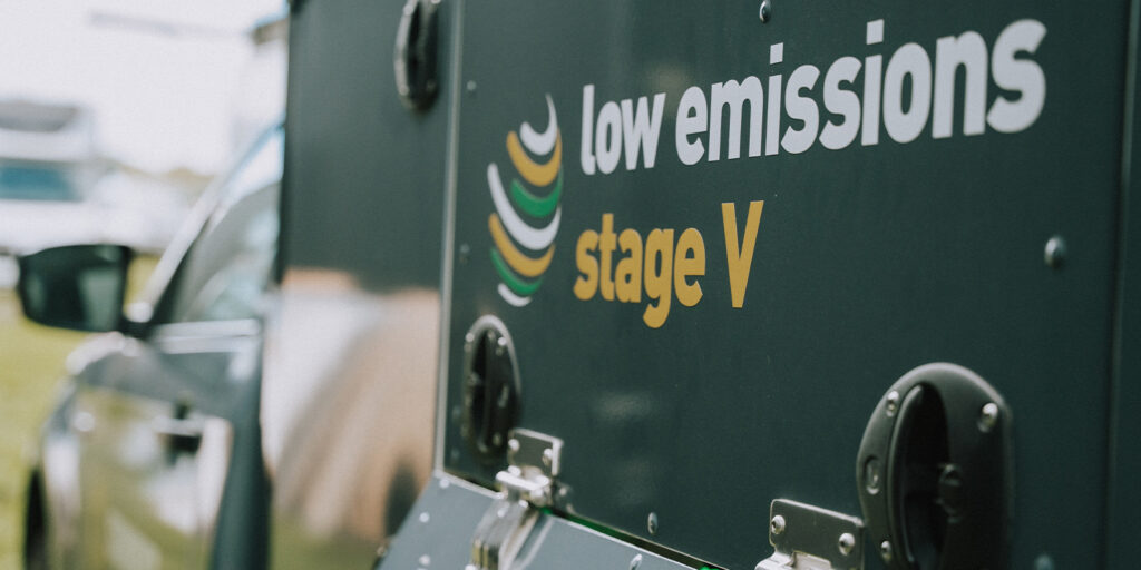 Close-up of a dark-coloured vehicle or equipment in the spotlight, displaying the text low emissions stage V with an abstract graphic, highlighting sustainability and compliance with environmental emission standards. A car is partially visible in the background.