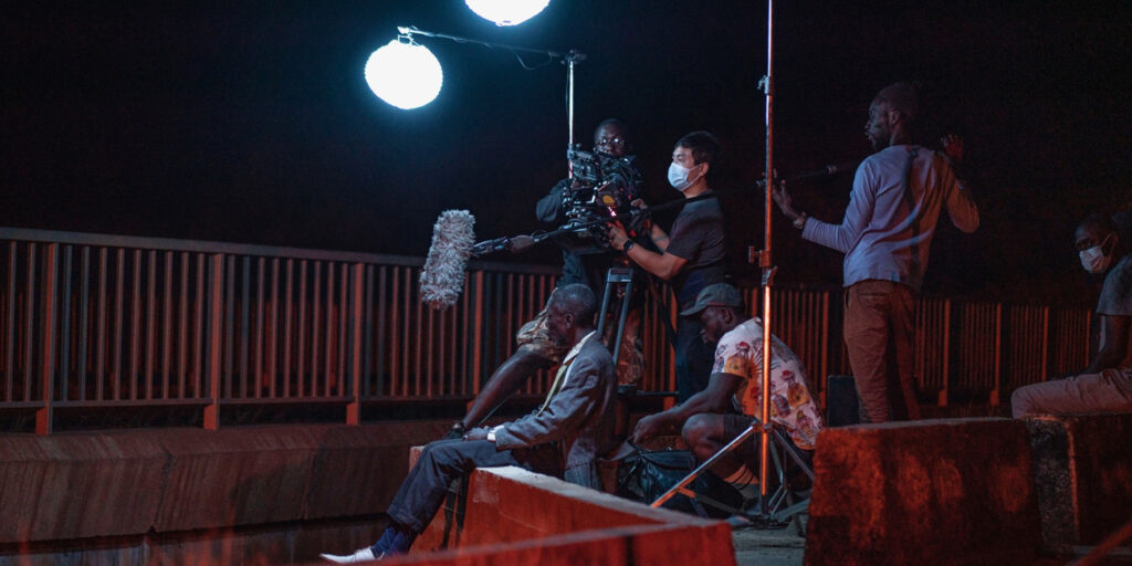 A film crew records at night with two large lights, a camera, and a boom microphone. Several people sit or stand near a metal railing, some wearing face masks. The outdoor set suggests efforts towards sustainability during production.