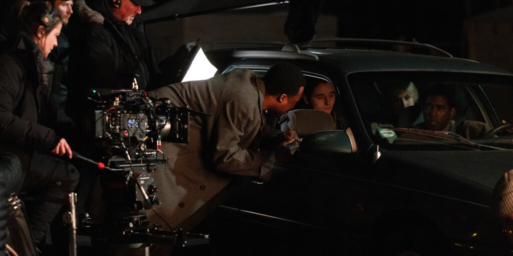 A film crew, led by Ula Pontikos BSC, shoots a night scene for The Man in My Basement, with an actor leaning into a car window. Visible camera and lighting equipment illuminate the set as crew members work nearby.