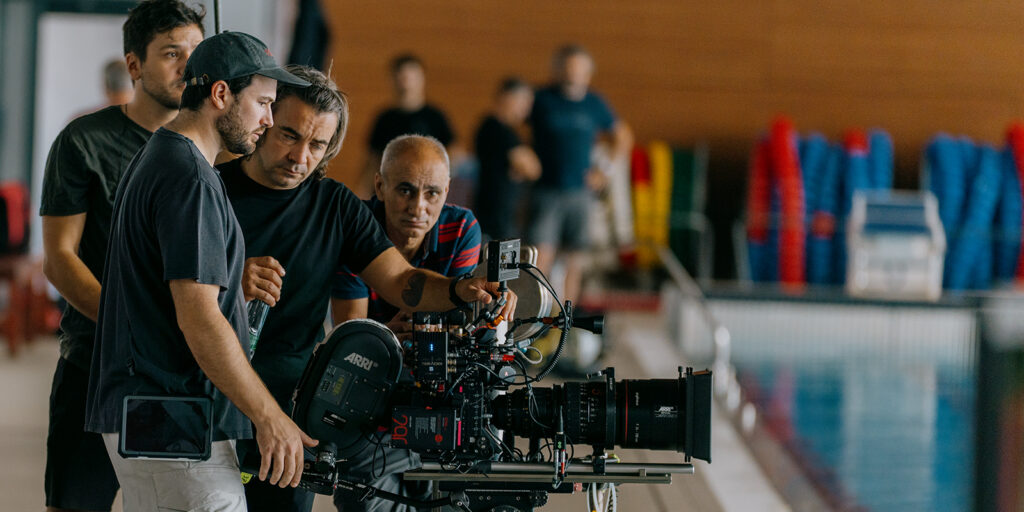 Several men, including Steven Breckon, stand by a swimming pool, focused on a professional film camera setup, possibly reviewing footage for The Plague. Colourful pool equipment is visible in the background.