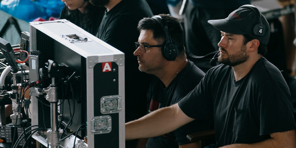 Two men in black shirts and caps sit at a workstation with large monitors and audio equipment, focused on their screens. One, Steven Breckon, wears headphones as they work behind the scenes on The Plague film or video production.