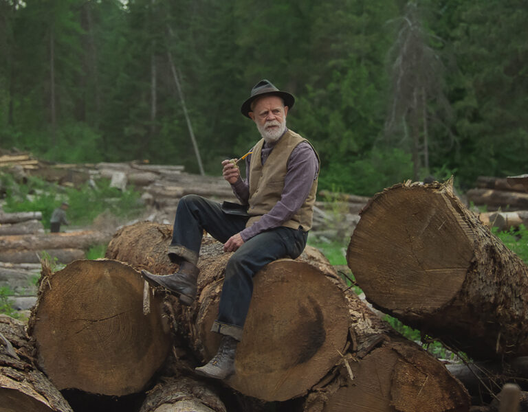 An older man in a waistcoat and hat sits on large cut tree logs, holding a pipe, whilst other men work with saws and axes around him in a forested logging area—an image of Train Dreams, captured in the style of Adolpho Veloso.