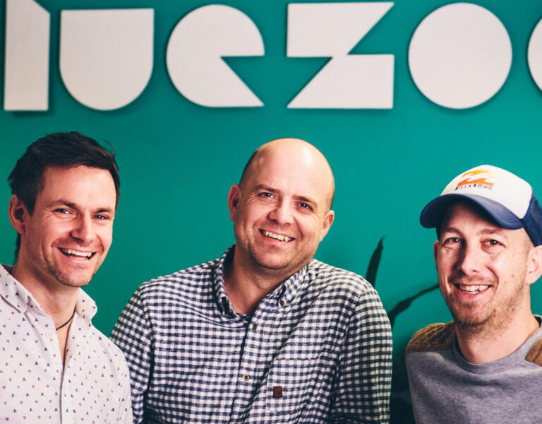 Three men smiling in front of a Blue Zoo logo