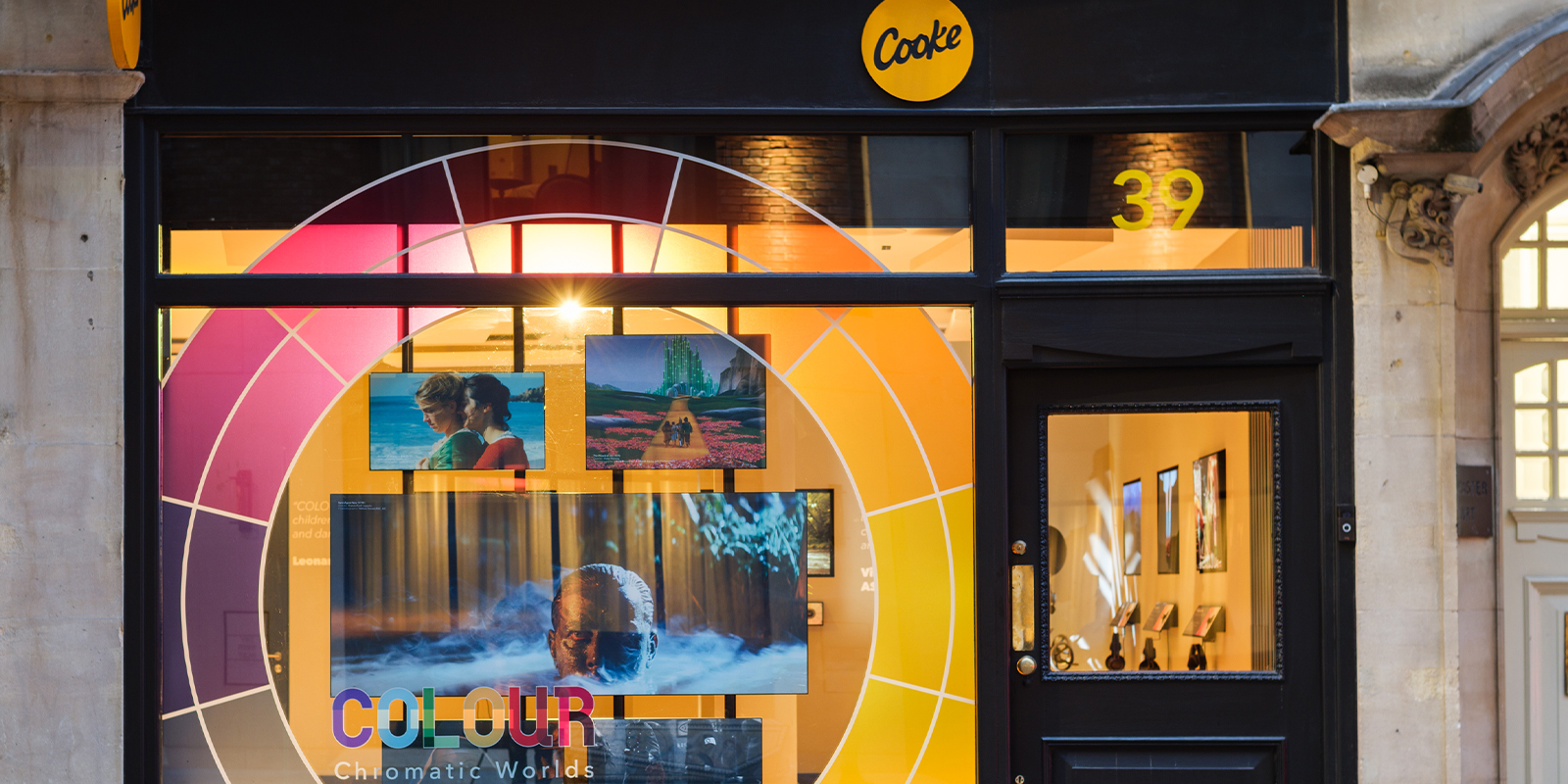 Shopfront with a large circular colour wheel graphic, “Cooke” logo above the door, and number 39 on the glass. Screens display various images for a Cooke Optics exhibition, with “COLOUR Chromatic Worlds” on the window.