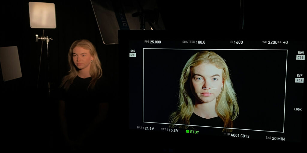 A woman with long blonde hair stands in front of a black background, surrounded by studio lights arranged for lighting for balance. Her face appears on a camera monitor in the foreground, framed for a close-up shot.